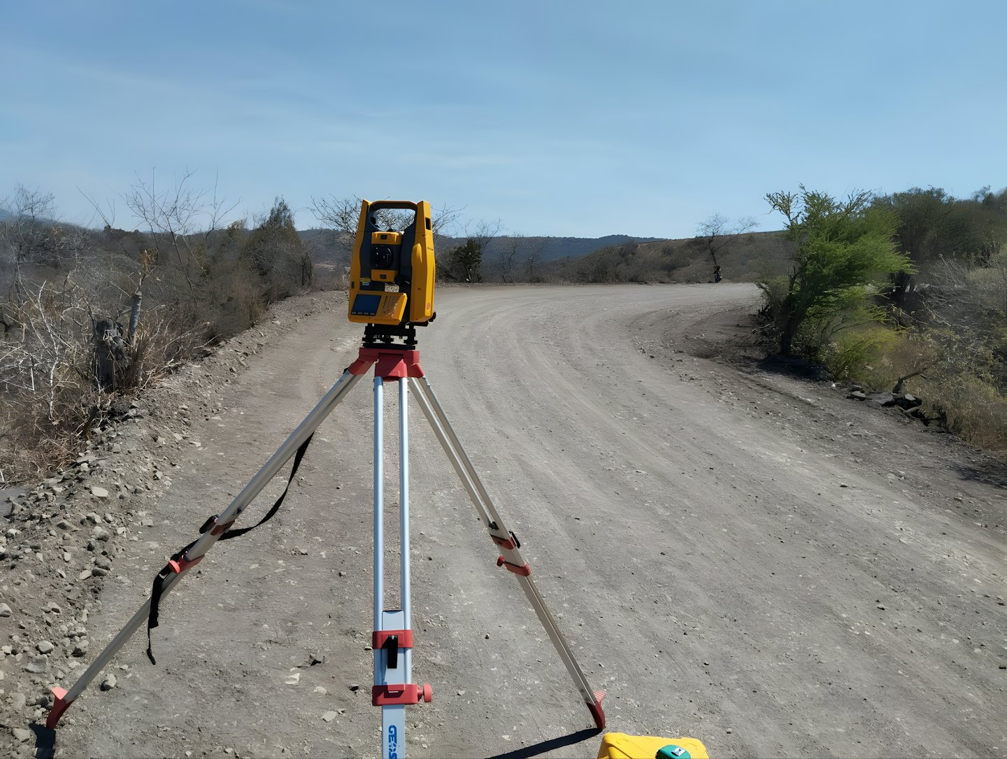 A yellow surveying total station stands on a tripod on a dirt road.